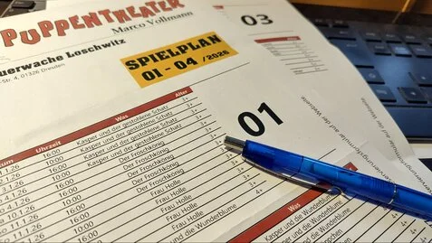 Eine Nahaufnahme einer Tabelle mit dem Puppentheaterspielplan, die auf einer Computertastatur liegt. Auf dem papier liegt ein blauer Kugelschreiber.